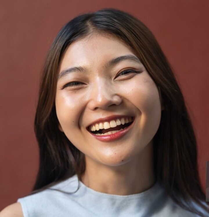 Radiant young woman with a contagious smile poses confidently in front of a warm red wall, exuding positivity and happiness. Young individuals with colored/textured backdrops + in outside environments concept.